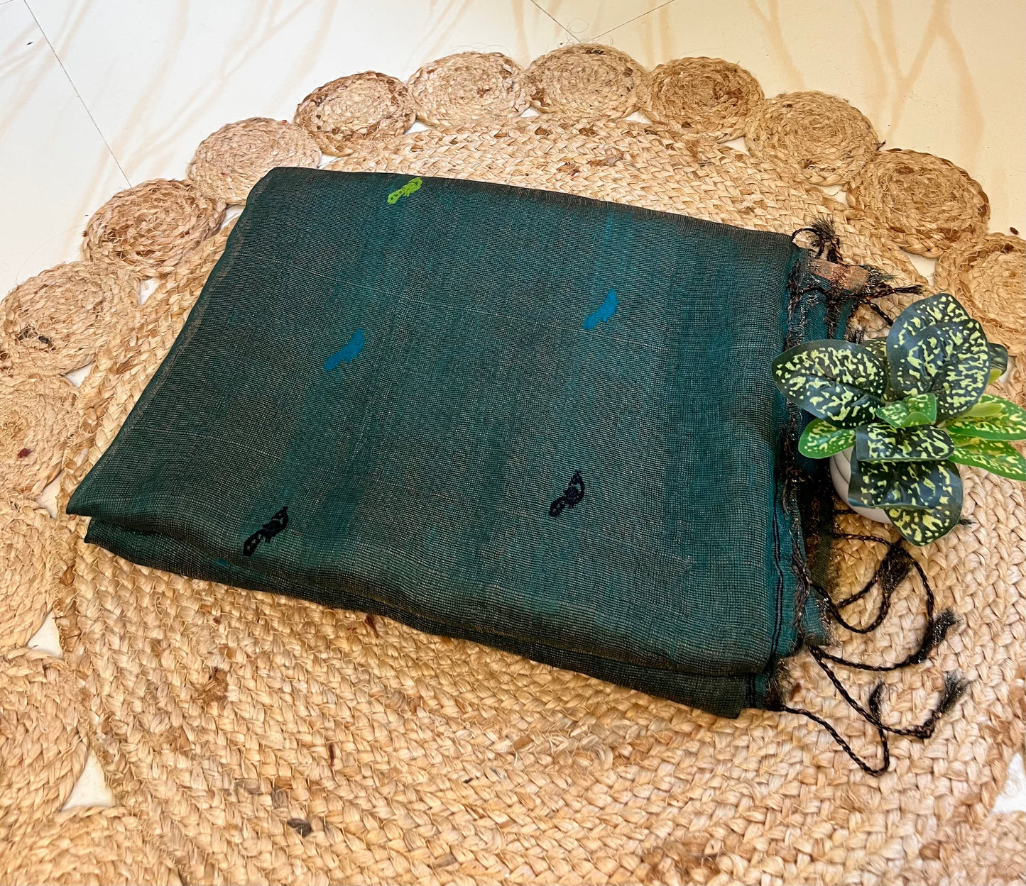 A folded dark green linen shawl with small embroidered motifs, displayed on a round woven jute placemat next to a small green plant.