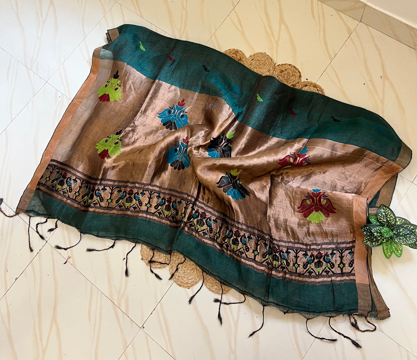 A draped silk saree or shawl in earthy brown and deep teal green, decorated with colorful floral embroidery and a patterned border, laid out on a light beige tiled floor.