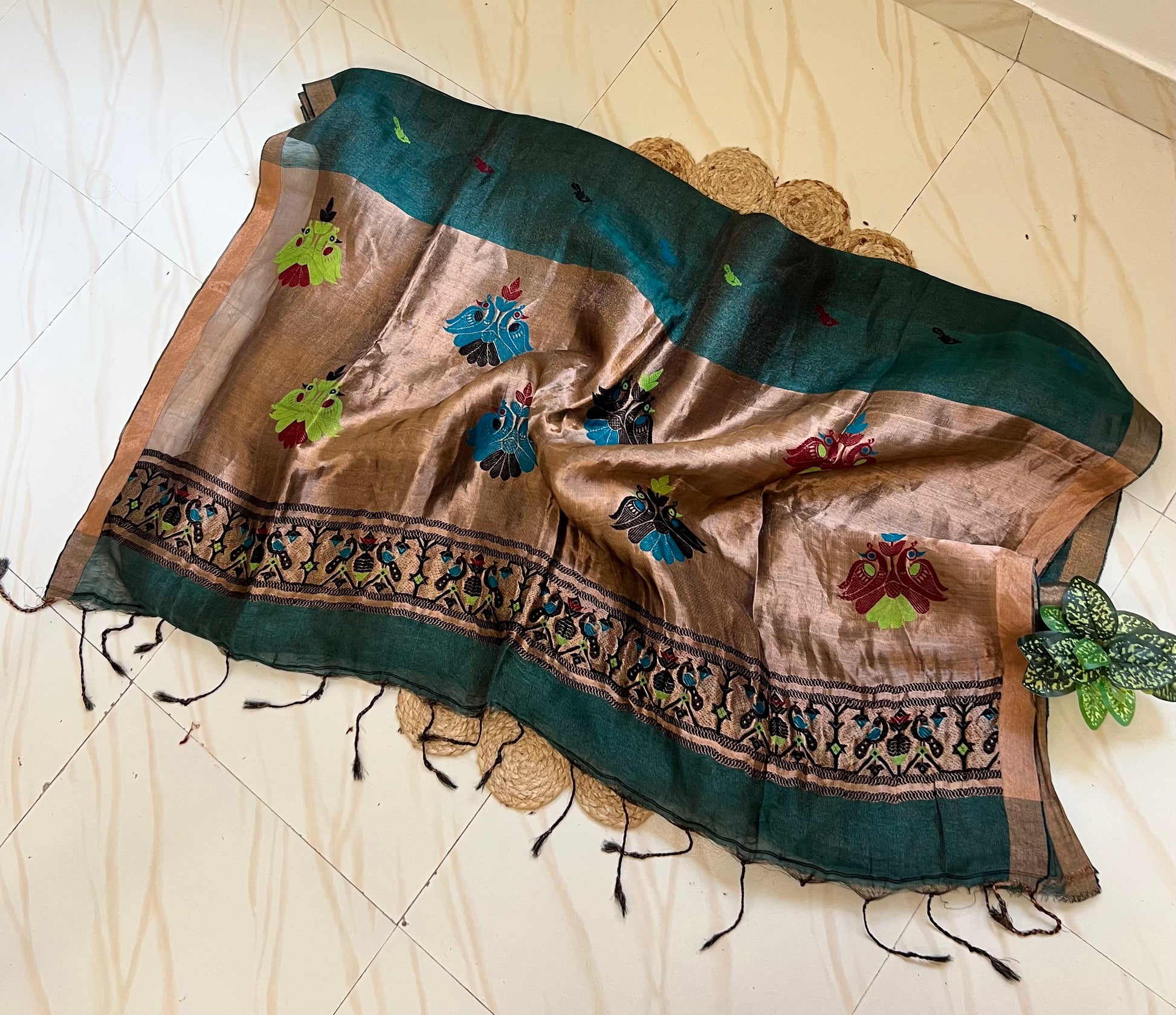 A draped silk saree or shawl in earthy brown and deep teal green, decorated with colorful floral embroidery and a patterned border, laid out on a light beige tiled floor.
