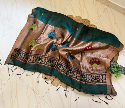 A draped silk saree or shawl in earthy brown and deep teal green, decorated with colorful floral embroidery and a patterned border, laid out on a light beige tiled floor.