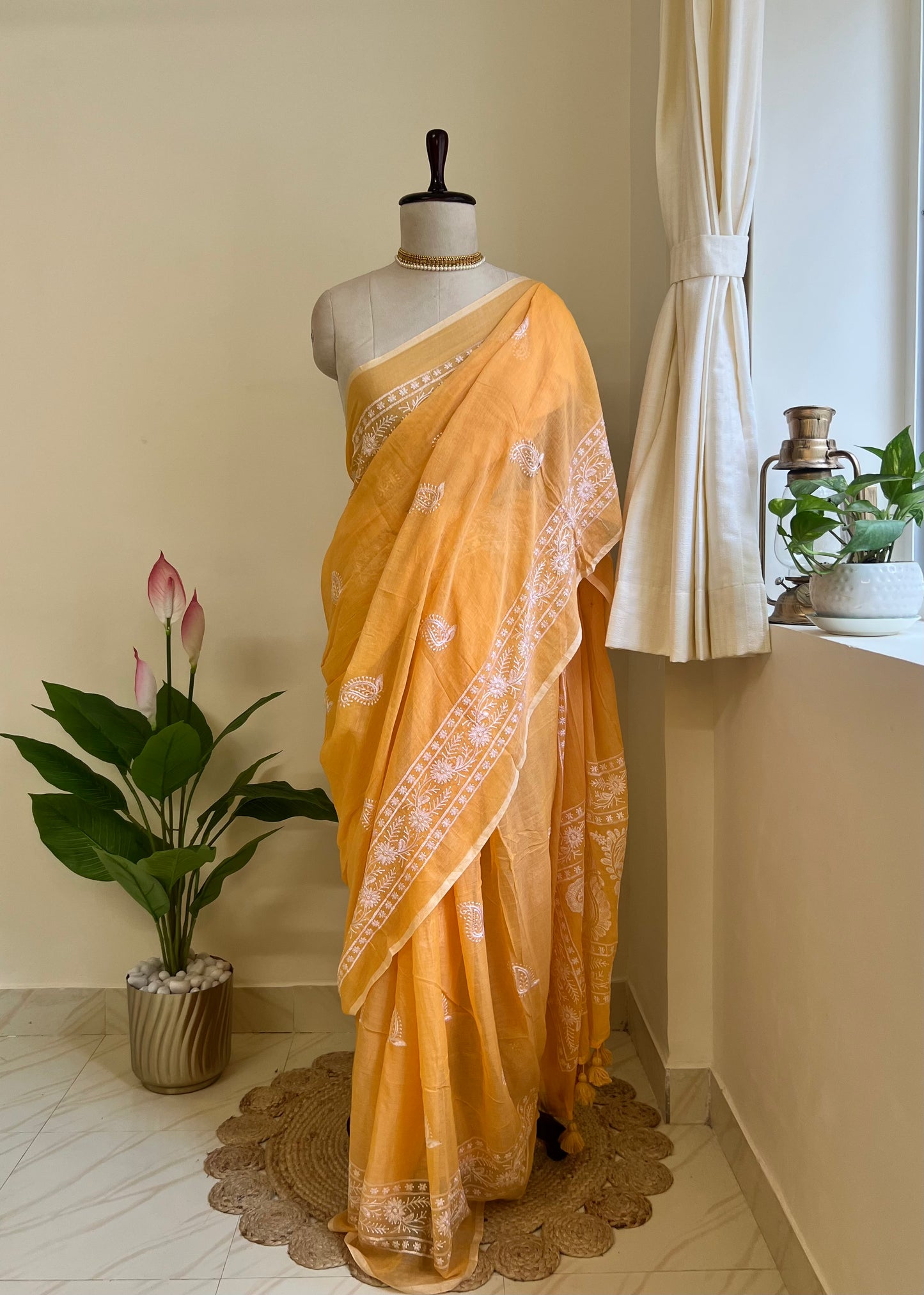 Marigold orange mul cotton saree with white chikankari-style embroidery, draped on a mannequin near a window with hanging curtains